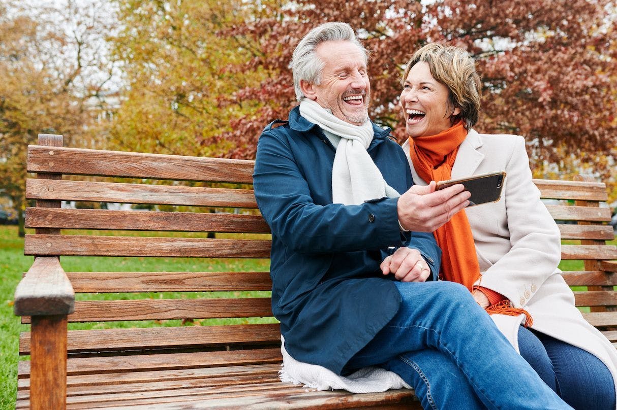 Older couple laughing at a phone, sitting on a bench